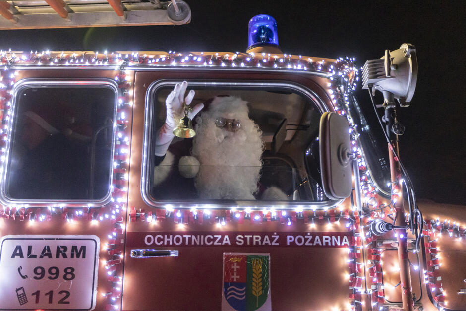 na zdjęciu mikołaj w biało czerwonym stroju z białą brodą, z dzwoneczkiem w ręce, jedzie czerwonym samochodem