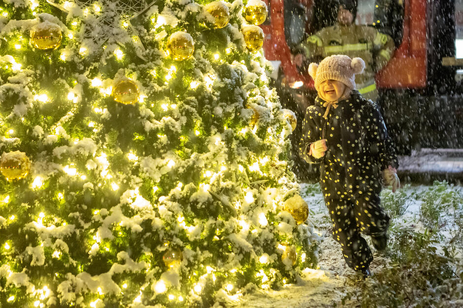 małe dziecko, prawdopodobnie około 2-3 lat, biegnie w kierunku dużej choinki pokrytej śniegiem. Dziecko nosi czarno-biały kombinezon i czapkę z futrzanym pomponem. Uśmiecha się i cieszy opadami śniegu. W tle zaparkowana jest czerwona ciężarówka strażacka 