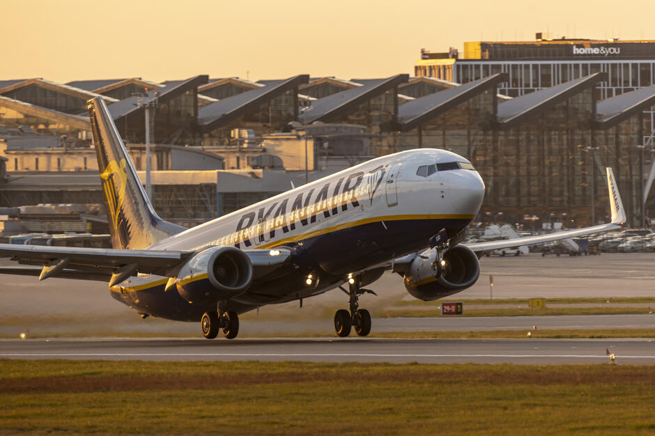 Samolot linii Ryanair unosi się od pasa startowego w ciepłym, złotym świetle zachodzącego słońca. W tle widać terminal lotniska z charakterystycznymi dachami oraz zaparkowanym sprzętem obsługi naziemnej. 