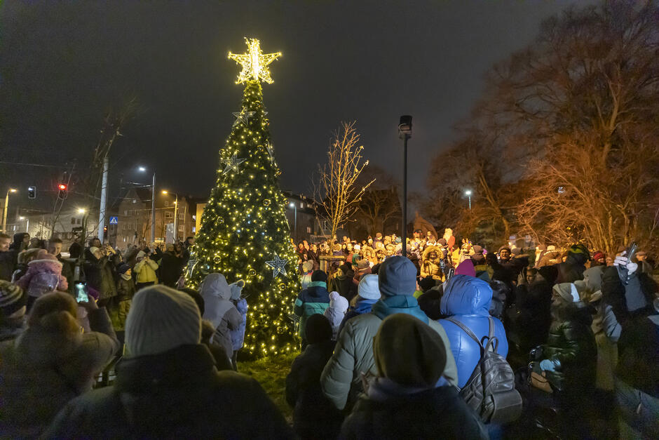 duża choinka w środku parku w nocy. Drzewo jest ozdobione kolorowymi światłami i gwiazdą na górze. Jest otoczone tłumem ludzi, niektórzy trzymają telefony do robienia zdjęć 