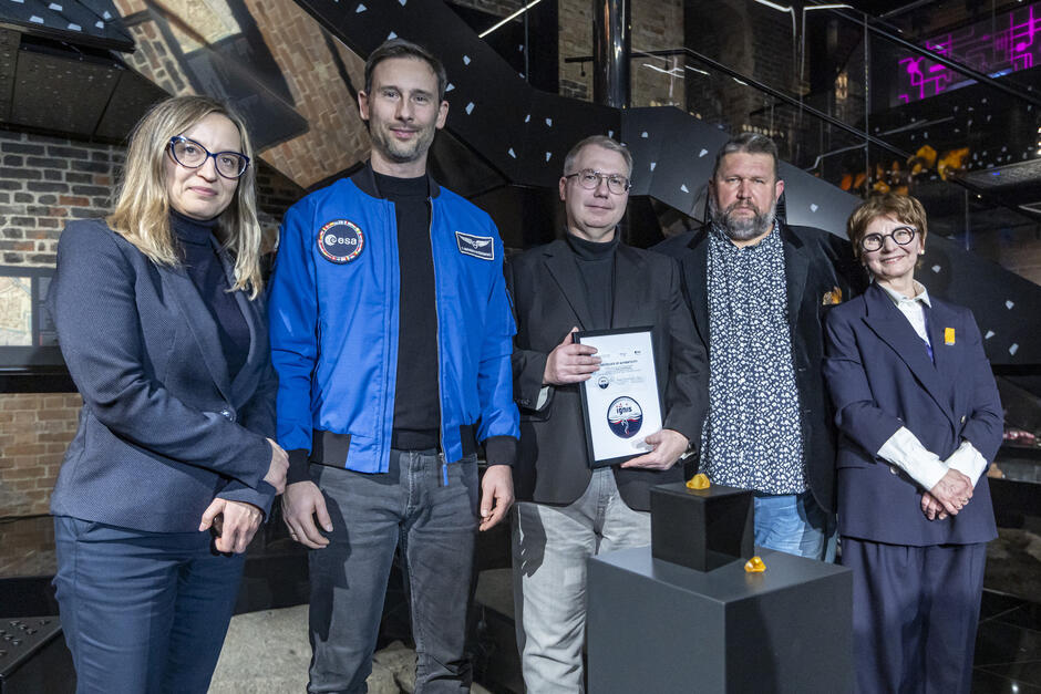 Dwie kobiety i trzech mężczyzn pozują do zdjęcia w Muzeum Bursztynu. Przed nimi na czarnym podeście leżą dwie bryłki bursztynu.