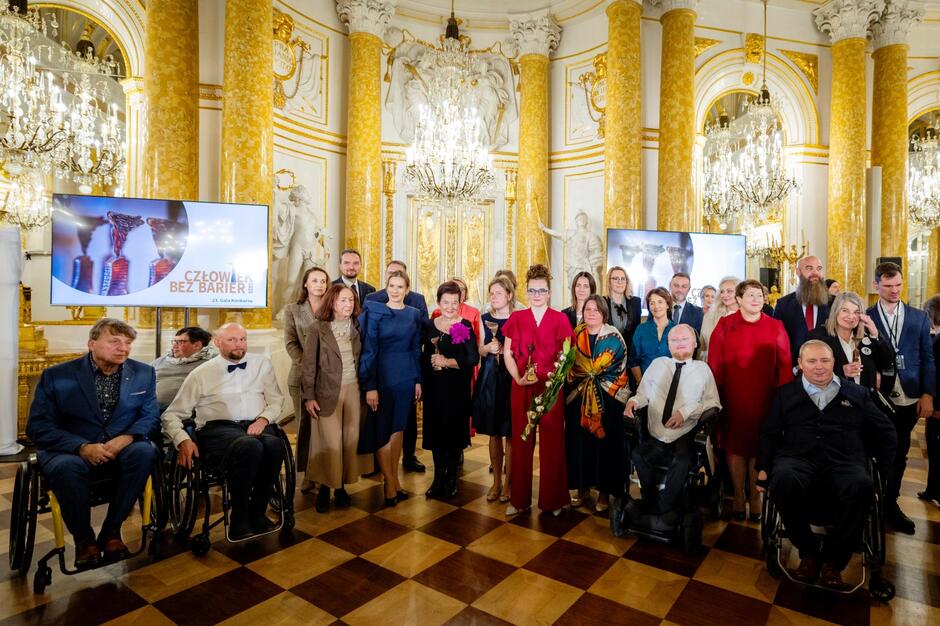 Grupa uczestników ceremonii pozuje do wspólnego zdjęcia w bogato zdobionej, pałacowej sali z kolumnami i kryształowymi żyrandolami. W tle widoczne są ekrany z identyfikacją wydarzenia, a wśród zgromadzonych znajdują się osoby trzymające statuetki oraz poruszające się na wózkach.
