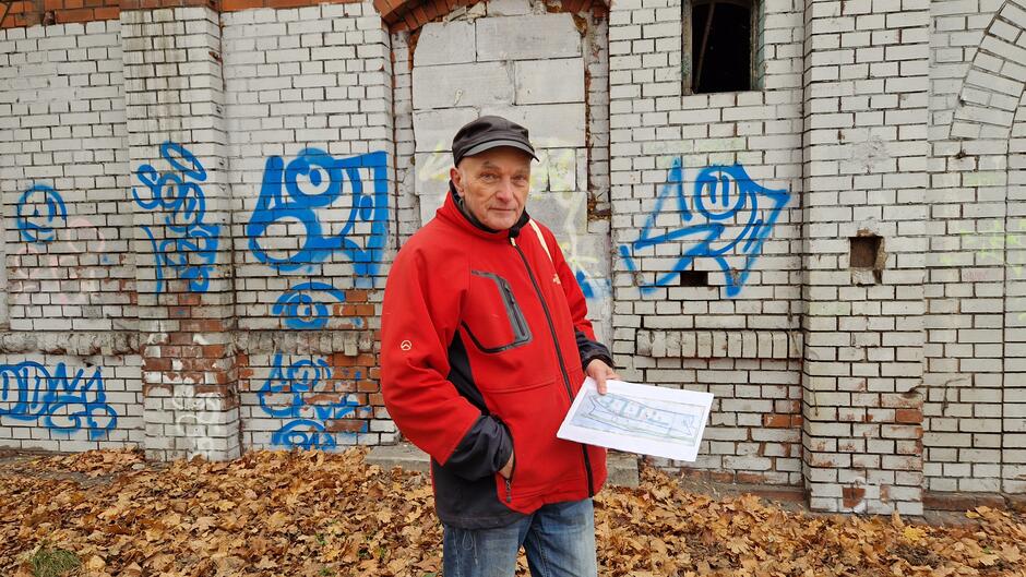 Starszy mężczyzna w czerwonej zimowej kurtce stoi na tle ceglanego budynku, pomalowanego na biało i zabrudzonego niebieskimi napisami grafitti