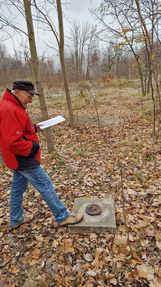 Starszy mężczyzna w czerwonej zimowej kurtce stawia nogę na wybetonowym kwadracie z metalowym okrągłym zamknięciem
