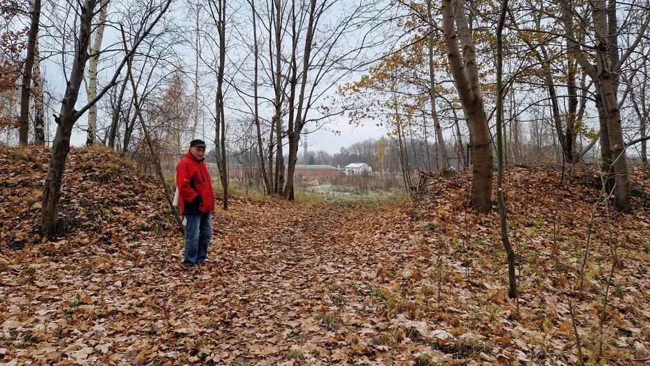 Mężczyzna w czerwonej zimowej kurtce wśród drzew i niskich wałów ziemnych pokrytych liśćmi, w tle widać Martwą Wisłę i Twierdzę Wisłoujście