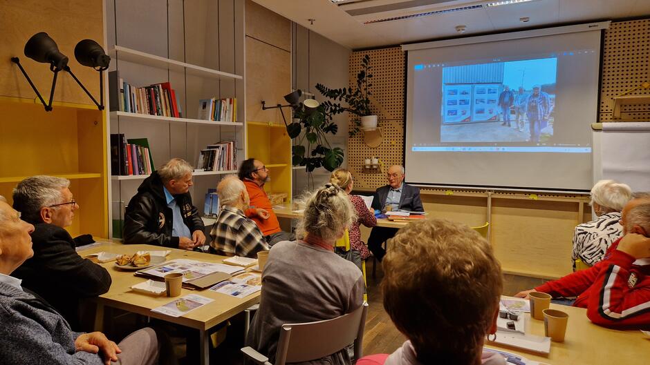 Widoczna grupa ludzi na zebraniu w niewielkiej sali. Za prowadzącym jest ekran z wyświetlaną przentacją