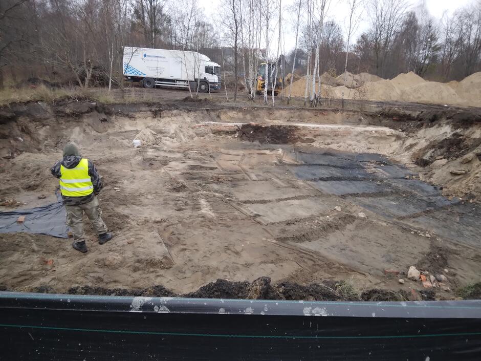Szeroki, ale dość płytki wykop ziemny wśród drzew. Na pierwszym planie widoczny człowiek z w kamizelce odblaskowej, z tyłu ciężarówka