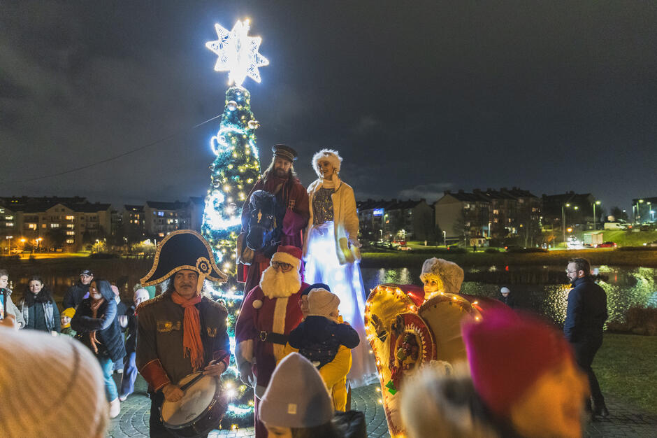 grupa ludzi zebranych wokół dużej choinki w parku wieczorem. Drzewo jest ozdobione kolorowymi światłami i gwiazdą na górze. W grupie jest sześć osób, trzech mężczyzn i dwie kobiety, ubrane w bajkowe kostiumy świąteczne. Jedna gra na bębnie,