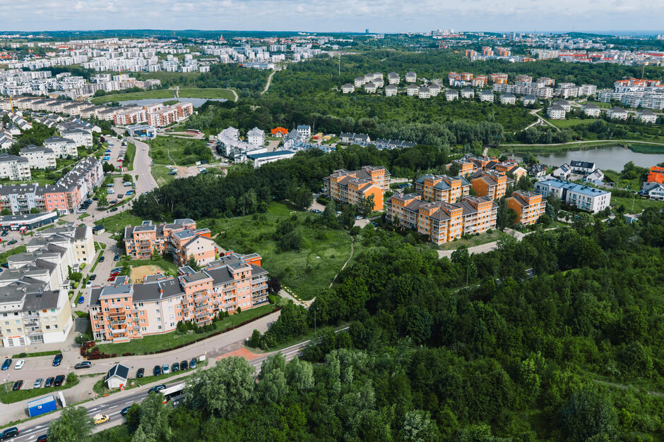 Panoramiczny widok ukazuje rozbudowane osiedla mieszkaniowe Gdańska otoczone dużymi terenami zielonymi i układem lokalnych dróg. W tle widoczne są kolejne skupiska zabudowy oraz zbiornik wodny, podkreślające dynamiczny rozwój tej części miasta.
