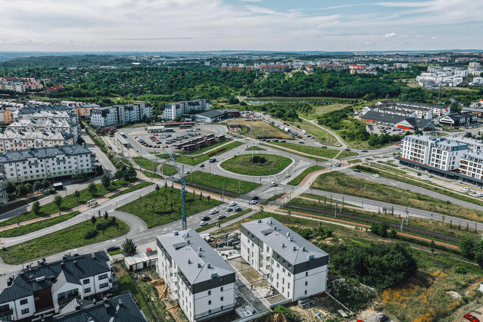 Zdjęcie przedstawia rozbudowany układ drogowy z dużym rondem otoczonym nową zabudową mieszkaniową oraz obiektami usługowymi. W tle widoczne są gęsto zalesione tereny i kolejne osiedla, tworząc dynamiczny miejski krajobraz tej części Gdańska.
