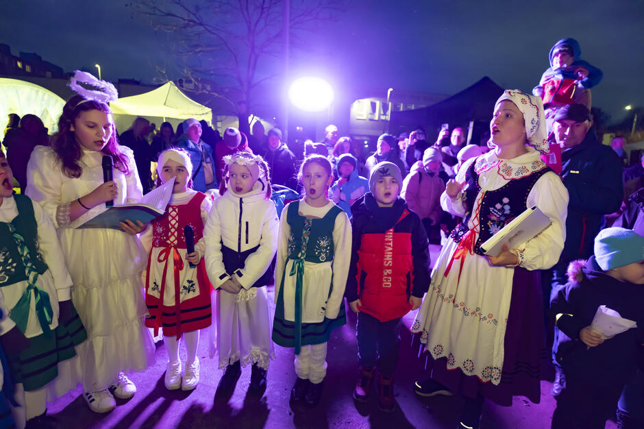 Grupa dzieci w strojach aniołów i regionalnych strojach ludowych śpiewa kolędy podczas plenerowego wydarzenia po zmroku. W tle widać zgromadzoną publiczność, namioty oraz sceniczne oświetlenie nadające wydarzeniu świąteczną oprawę.
