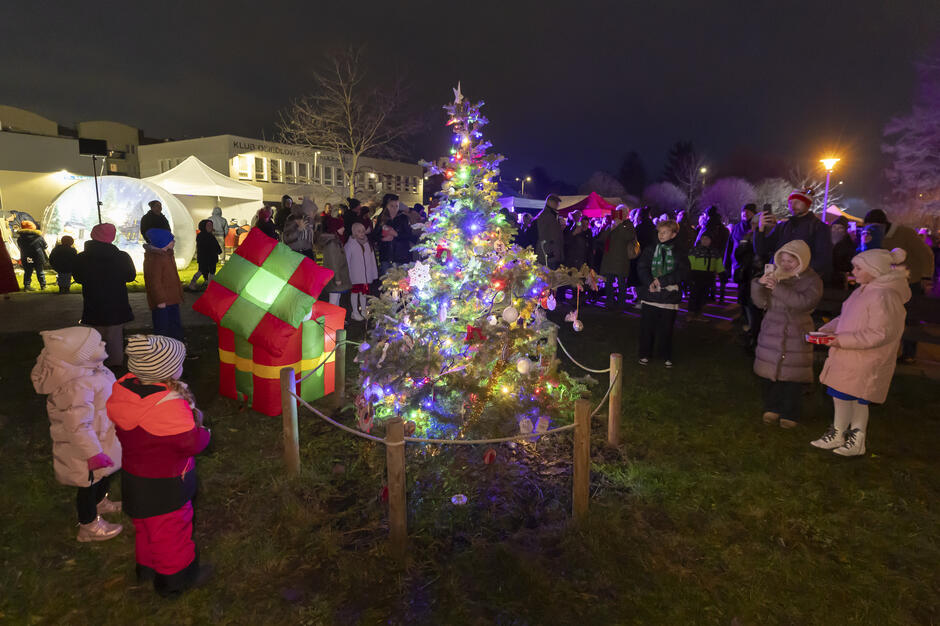 Uczestnicy wydarzenia gromadzą się wokół niewielkiej, udekorowanej choinki rozświetlonej kolorowymi lampkami. Wokół stoją dzieci i dorośli w zimowych ubraniach, a w tle widoczne są namioty oraz świąteczna iluminacja.
