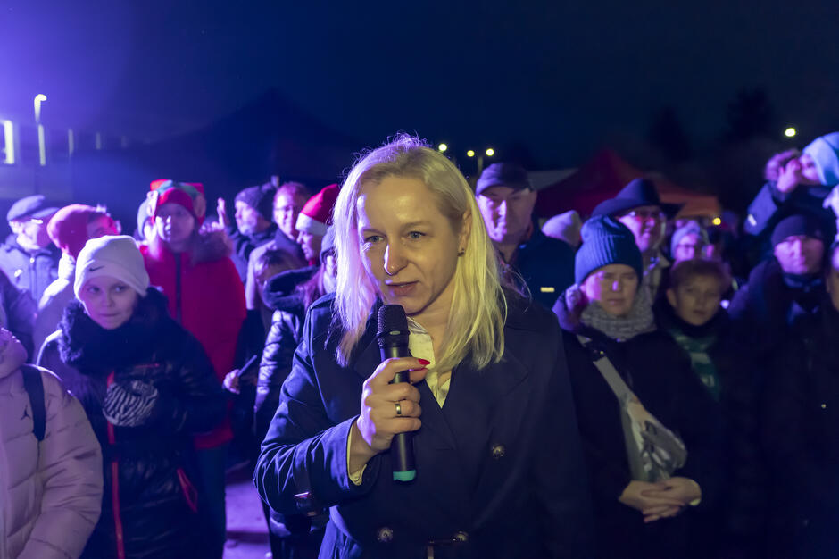 Kobieta przemawia do zgromadzonych uczestników wydarzenia, trzymając mikrofon i stojąc wśród tłumu na świeżym powietrzu po zmroku. W tle widoczna jest publiczność ubrana w zimowe kurtki i czapki, oświetlona światłem scenicznym.
