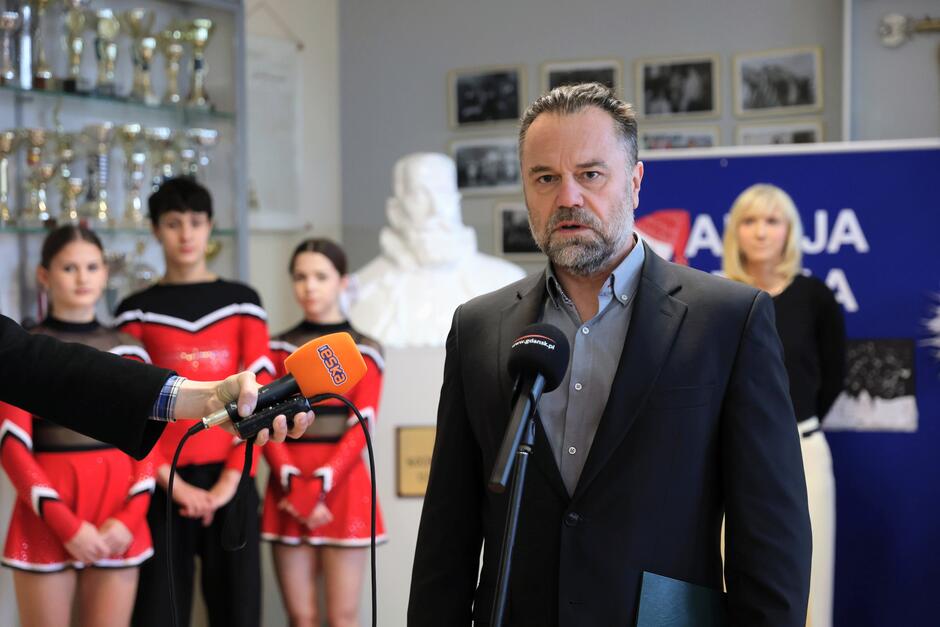 na zdjęciu mężczyzna w średnim wieku, w czarnej marynarce, mówi do mikrofonu dziennikarskiego