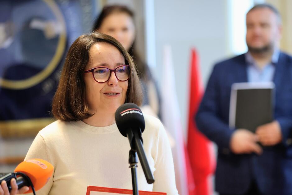 na zdjęciu kobieta w średnim wieku, szatynka w okularach, mówi do mikrofonu