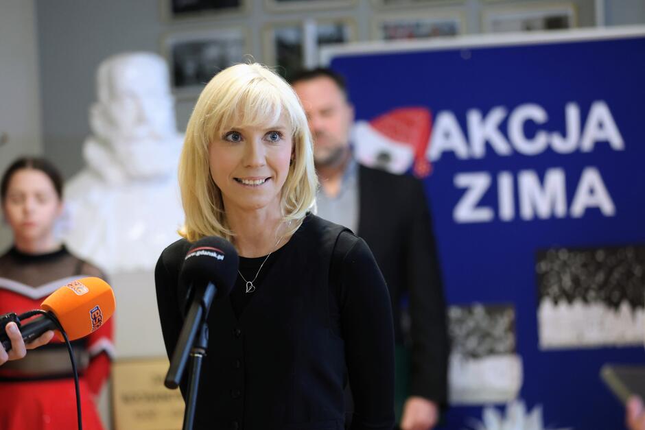 na zdjęciu szczupła blondynka, mówi do mikrofonu dziennikarskiego