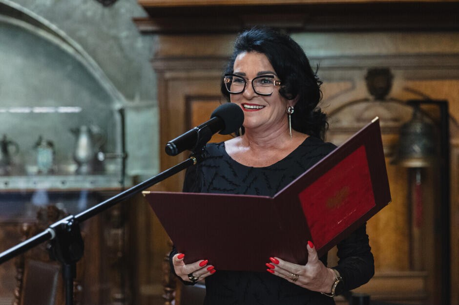 Kobieta stoi przy mównicy i czyta z otwartej teczki, przemawiając do mikrofonu w eleganckim, zabytkowym wnętrzu. Jej uśmiech i formalny strój podkreślają uroczysty charakter wydarzenia. 