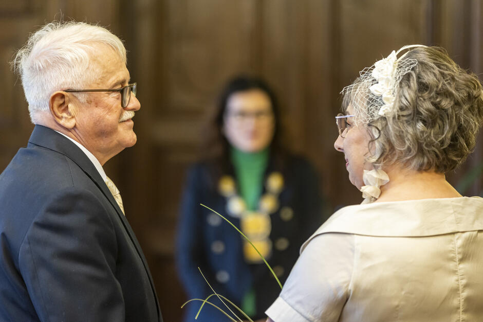 Starsza para stoi naprzeciw siebie w trakcie uroczystej ceremonii, patrząc sobie w oczy w eleganckim, drewnianym wnętrzu. W tle widoczna jest postać urzędniczki z insygniami, co podkreśla oficjalny charakter wydarzenia. 