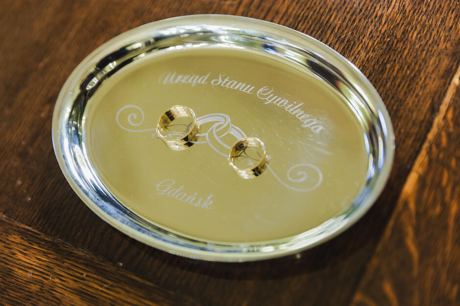 Na ozdobnej, jasnej tacke leżą dwie złote obrączki ślubne ułożone obok siebie. Grawer z nazwą urzędu i miasta podkreśla oficjalny charakter ceremonii.
