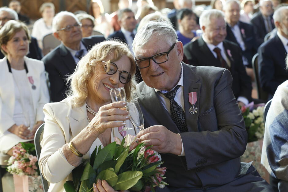 Starsza para siedzi obok siebie podczas uroczystości, wznosząc toast kieliszkami i uśmiechając się w ciepłej, odświętnej atmosferze. Wokół nich widoczni są inni uczestnicy wydarzenia, a kwiaty i odznaczenia podkreślają podniosły charakter spotkania. 