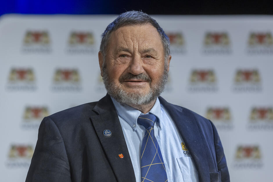 Na zdjęciu widoczny jest starszy mężczyzna z siwą brodą, ubrany w ciemną marynarkę, jasną koszulę i krawat, stojący na tle ścianki z powtarzającymi się logotypami wydarzenia. Portret ma charakter oficjalny i został wykonany podczas uroczystości lub gali, o czym świadczy elegancki strój oraz przypinki na klapie marynarki. 