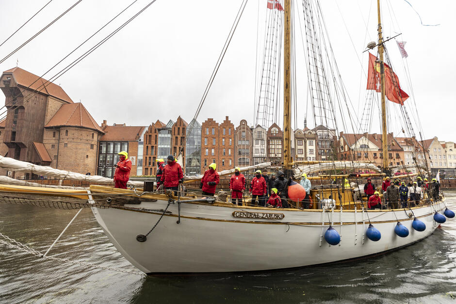 Na zdjęciu widoczny jest żaglowiec „Generał Zaruski” z załogą stojącą na pokładzie, ubranej w czerwone sztormiaki, podczas postoju na wodzie w miejskiej scenerii portowej. W tle widać historyczną zabudowę nabrzeża, a całość przedstawia moment oficjalnego wejścia lub powitania jednostki w porcie. 