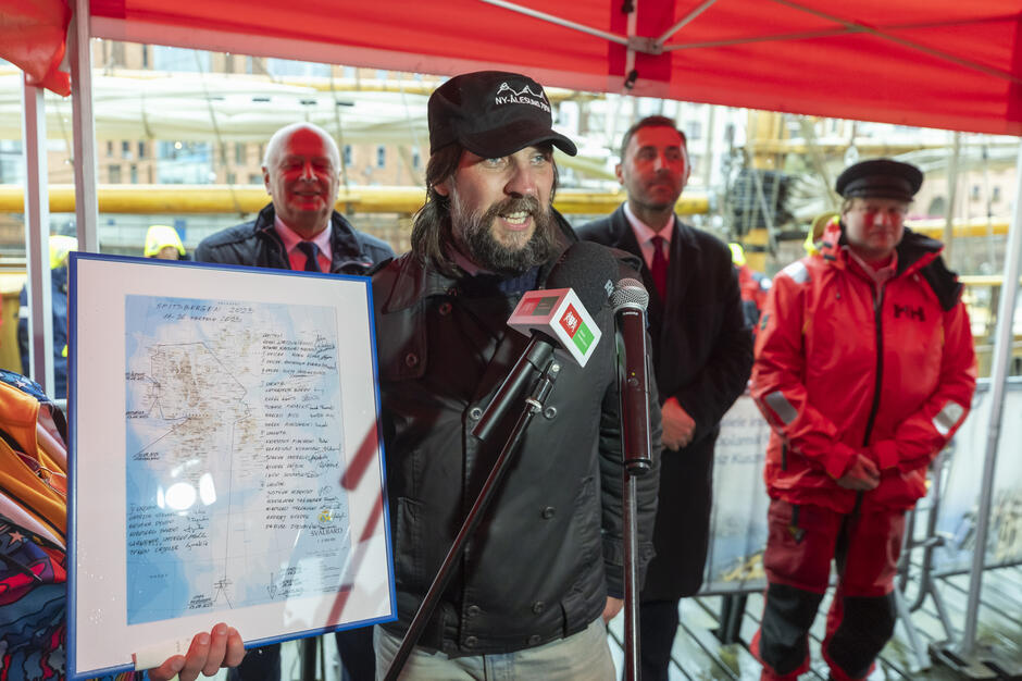 Na zdjęciu widoczny jest mężczyzna stojący pod namiotem, przemawiający do mikrofonów i trzymający w ręku oprawioną mapę lub dokument z odręcznymi zapiskami. W tle znajdują się inni uczestnicy wydarzenia, w tym osoby w strojach sztormowych, co wskazuje na oficjalne powitanie lub podsumowanie rejsu w portowej scenerii.
