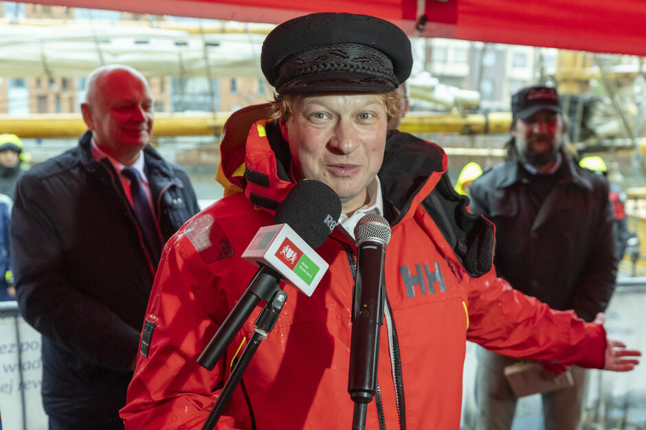 Na zdjęciu widoczna jest osoba w czerwonym sztormiaku i marynarskiej czapce, przemawzyżająca do mikrofonów podczas wydarzenia zorganizowanego pod namiotem w porcie. W tle znajdują się inni uczestnicy uroczystości oraz elementy infrastruktury żaglowca, co wskazuje na oficjalne powitanie lub wystąpienie po rejsie.
