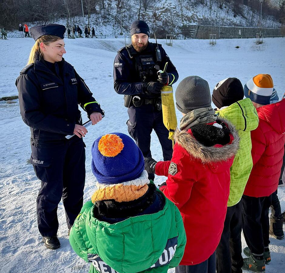 Dwoje funkcjonariuszy stoi na śniegu przed grupą dzieci. W rękach trzymają opaski odblaskowe.