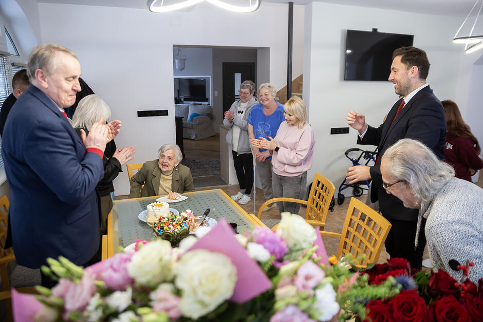 Przy stole siedzi stara kobieta, wokół stoi kilka osób i śpiewa, wszyscy się śmieją, niektórzy bija brawo.