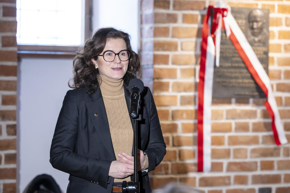 Na zdjęciu widoczna jest kobieta stojąca przy mikrofonie i przemawiająca w ceglanej przestrzeni wnętrza. W tle znajduje się pamiątkowa tablica z biało-czerwoną wstęgą, zamontowana na ścianie.
