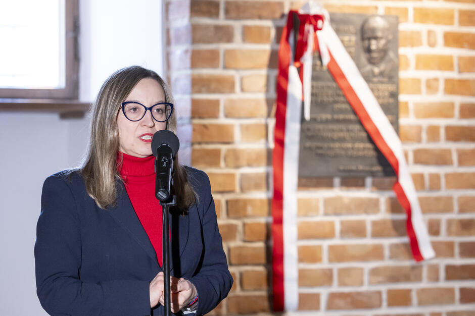 Na zdjęciu widoczna jest kobieta stojąca przy mikrofonie i przemawiająca w ceglanej przestrzeni wnętrza. W tle znajduje się pamiątkowa tablica z biało-czerwoną wstęgą, zamontowana na ścianie.
