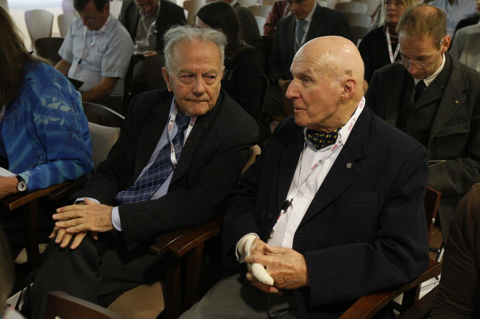 Na zdjęciu widoczni są dwaj starsi mężczyźni siedzący obok siebie na sali konferencyjnej i prowadzący rozmowę w trakcie wydarzenia. W tle znajdują się inni uczestnicy spotkania zajmujący miejsca na widowni.
