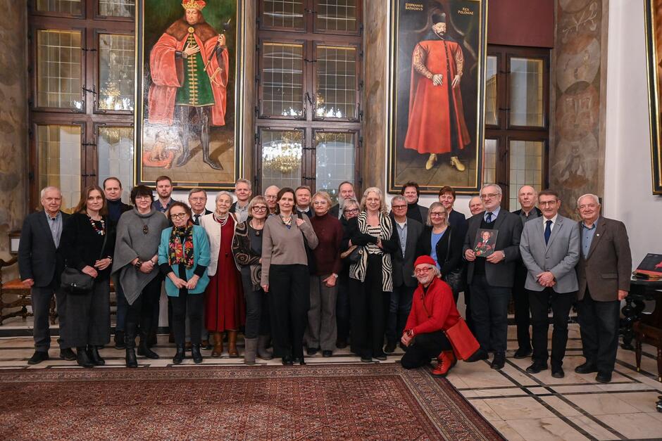 Grupowe zdjęcie przedstawia uczestników spotkania zgromadzonych w reprezentacyjnej sali z historycznymi portretami władców zawieszonymi na ścianach. Osoby pozują wspólnie, stojąc i klęcząc w jednym rzędzie, w eleganckim wnętrzu o charakterze pałacowym.
