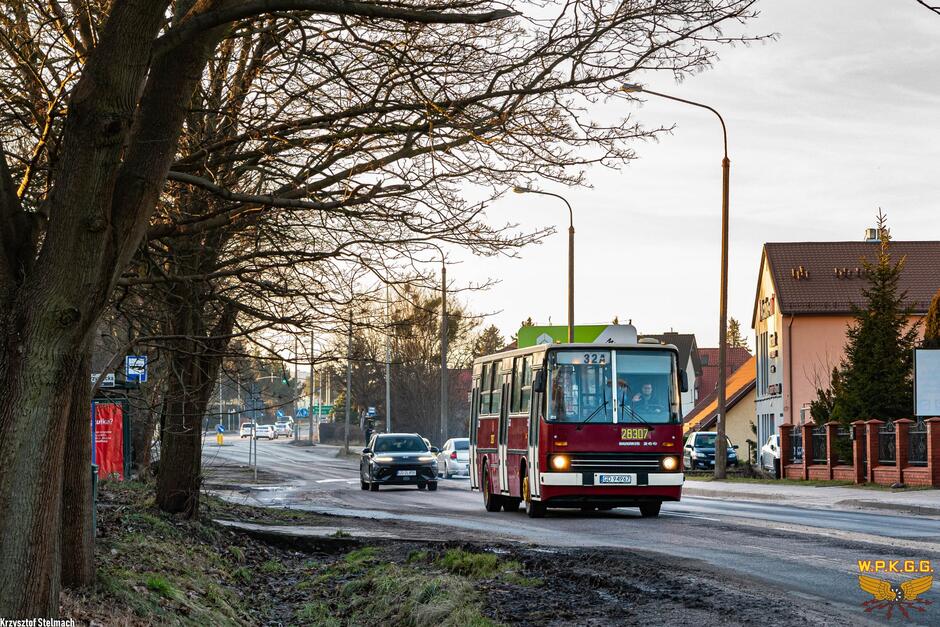 Zabytkowy autobus jedzie ulicą wśród niskiej zabudowy mieszkaniowej i bezlistnych drzew, w świetle niskiego, popołudniowego słońca. Na jezdni widoczne są także inne samochody, a całość tworzy spokojną, podmiejską scenerię.
