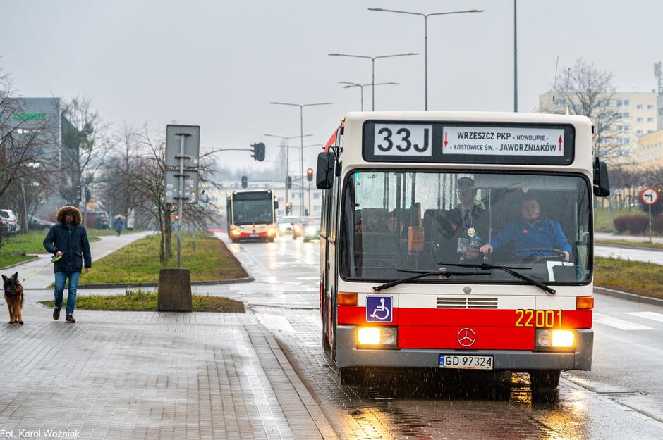 Autobus linii 33J stoi na przystanku przy mokrej jezdni, a przez przednią szybę widoczny jest kierowca i pasażerowie. W tle widać ruch uliczny, kolejny autobus oraz przechodnia z psem, co podkreśla codzienny, miejski charakter sceny.
