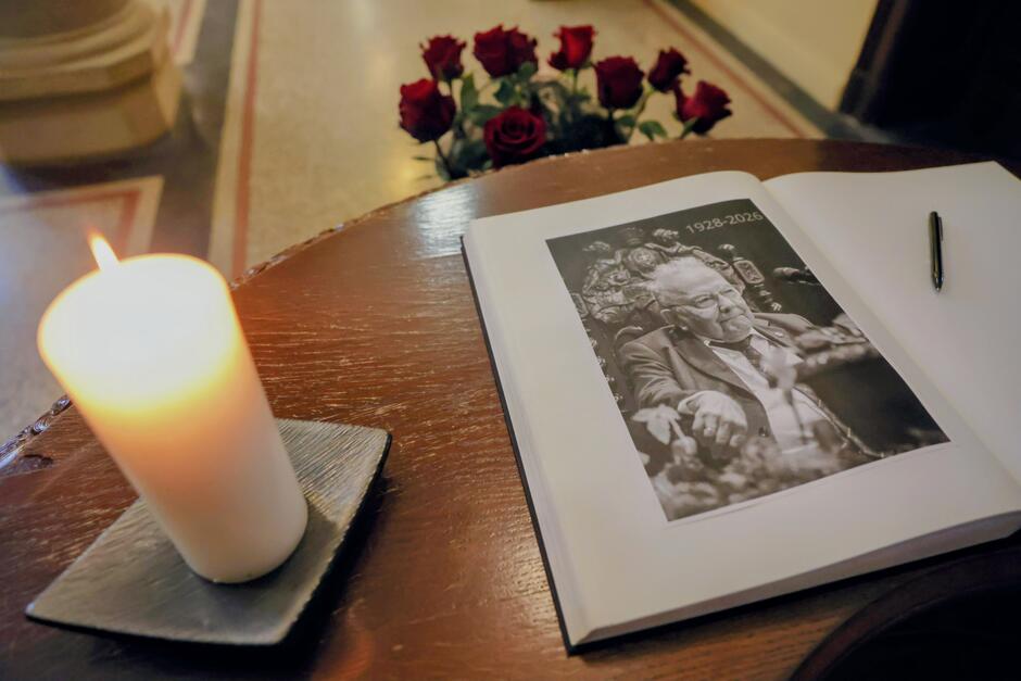 Na drewnianym pulpicie spoczywa księga pamiątkowa otwarta na stronie z czarno-białym portretem mężczyzny oraz datami 1928–2026, obok której leży długopis. Obok księgi pali się biała świeca, a w tle widoczny jest bukiet czerwonych róż, tworzący nastrojową, żałobną kompozycję.
