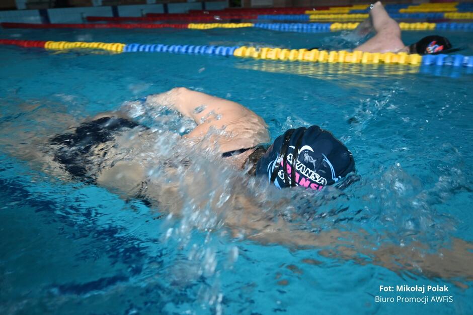 kobieta płynie w basenie pływackim, głowa w czepku jest pod wodą.