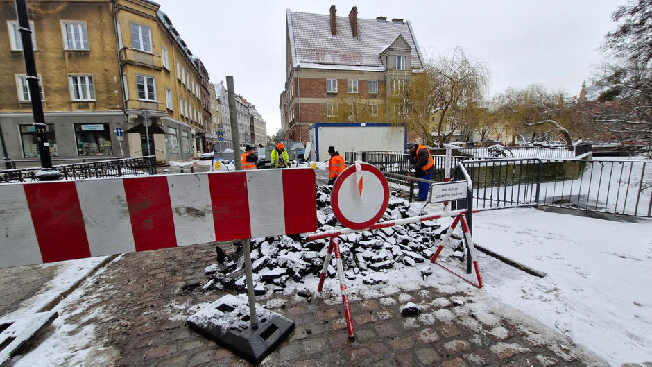 na zdjęciu widać znaki drogowe informujące o zamknięciu ulicy dla ruchu, w tle rozkopany mostek i dalej kilka kamienic