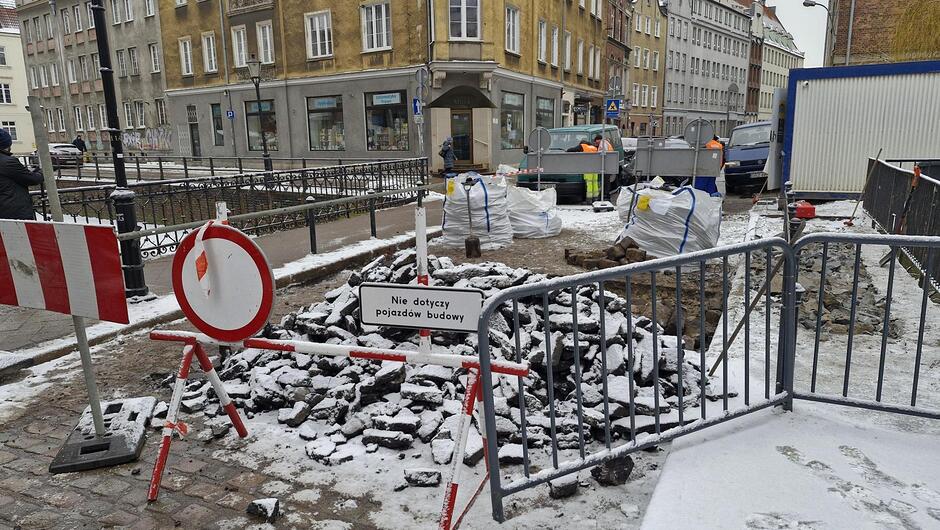 na zdjęciu widać zdjęty bruk z nawierzchni ulicy, wszystko pokryte jest śniegiem, w tle kilka kamienic