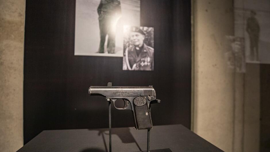 pistolet z czasów II wojny światowej na wystawie w ciemnym otoczeniu, w tle zdjęcia mjr. Sucharskiego