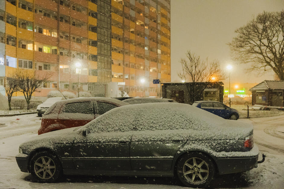 na pierwszym planie zaśnieżony samochód osobowy w ciemnym kolorze, obok stoi kilka innych samochodów, w tle ośnieżony wysoki blok mieszkalny