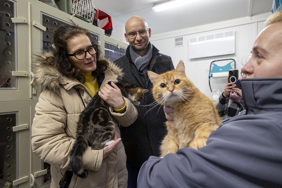 Dwie kobiety trzymają na rękach koty, obok stoi mężczyzna, w tle klatki dla kotów w schronisku dla bezdomnych zwierząt.