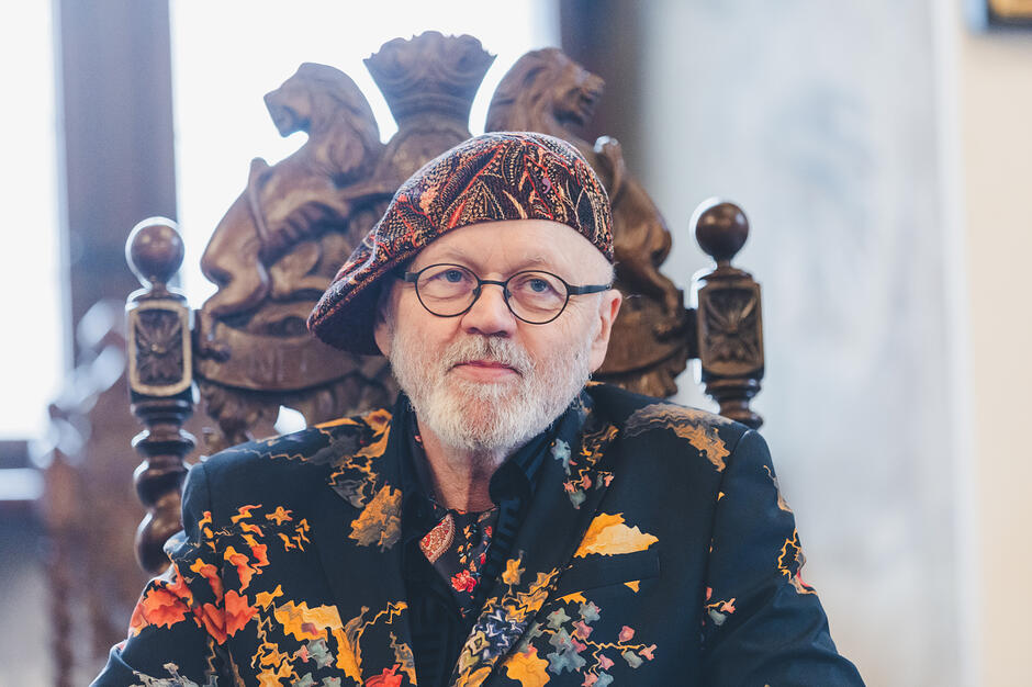 Mężczyzna w kolorowym stroju i berecie siedzi na rzeźbionym krześle gdańskim w reprezentacyjnej sali.