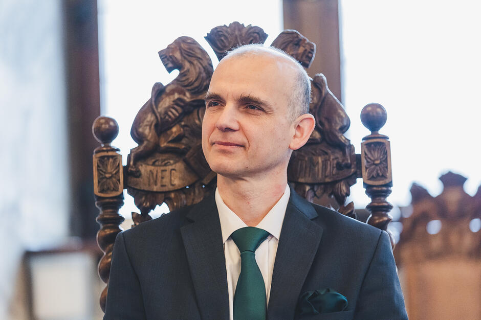 Mężczyzna w garniturze siedzi na rzeźbionym krześle gdańskim w reprezentacyjnej sali.