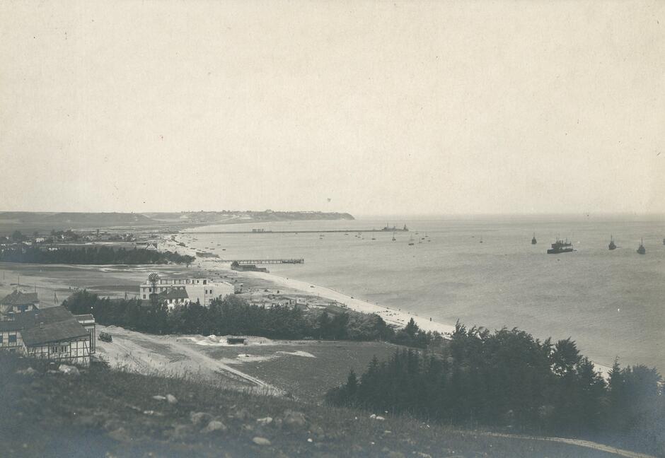 Czarnobiałe zdjęcie, widok na nadmorską wioskę z góry. Widoczne nieliczne domy, molo i łodzie na wodzie.