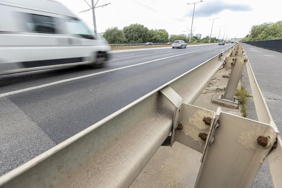 Na zdjęciu widać wielopasmową drogę z wyraźnie widocznymi, stalowymi barierami energochłonnymi na pierwszym planie. Po lewej stronie jezdni porusza się biały samochód dostawczy, a w oddali widać inne pojazdy oraz drzewa rosnące wzdłuż trasy