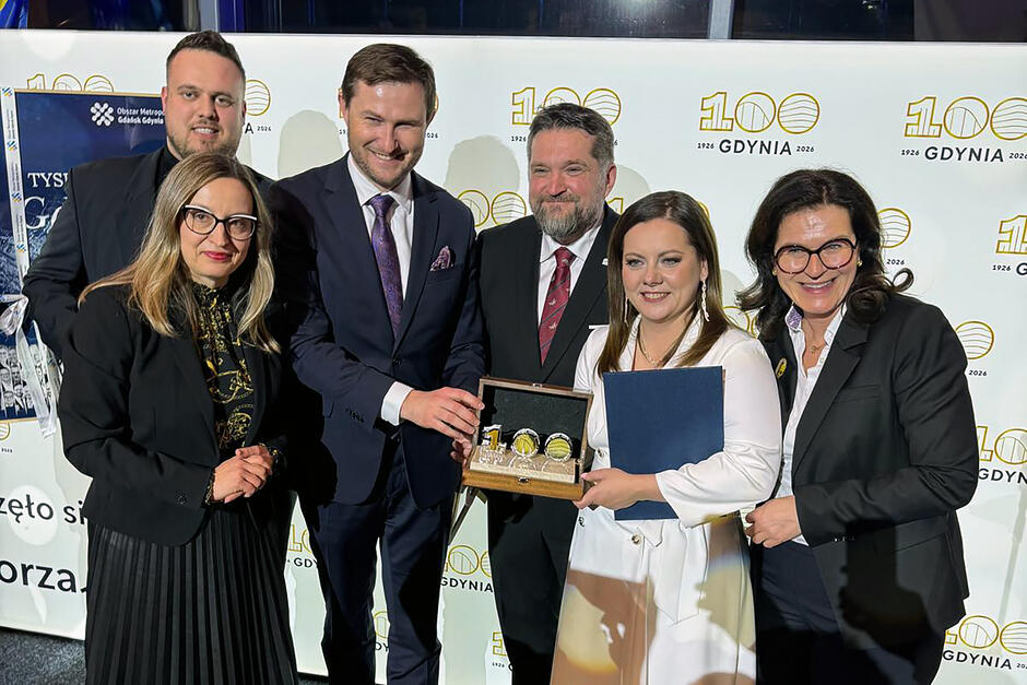 Grupa sześciu osób - trzy kobiety i trzech mężczyzn pozuje do zdjęcia na tle ścianki z napisem 100 lat gdyni . Ludzie uśmiechają się i pokazują statuetkę z napisem 100.