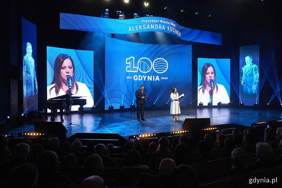 Kobieta i mężczyzna na scenie w teatrze, kobieta przemawia do mikrofonu, w tle ekran z napisem 100 lat Gdyni .