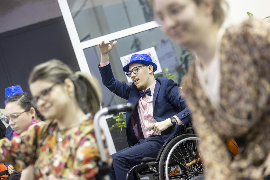 Na sali siedzi kilka osób, wśród nich mężczyzna na wózku inwalidzkim w garniturze, muszce pod szyja i kapeluszu.